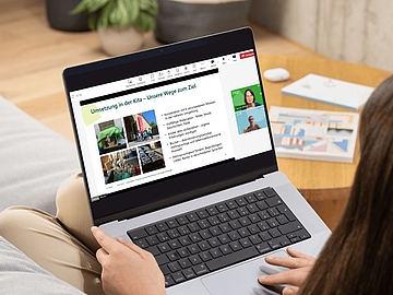 Eine Frau sitzt auf einem Sofa und schaut sich auf ihrem Laptop eine Online-Präsentation an. Auf dem Bildschirm ist eine Folie mit dem Titel „Umsetzung in der Kita – Unsere Wege zum Ziel“ zu sehen, die Fotos von Kita-Aktivitäten und eine Aufzählung von Maßnahmen zeigt. Rechts daneben sind zwei Vortragende in einem Videoanruf eingeblendet. 